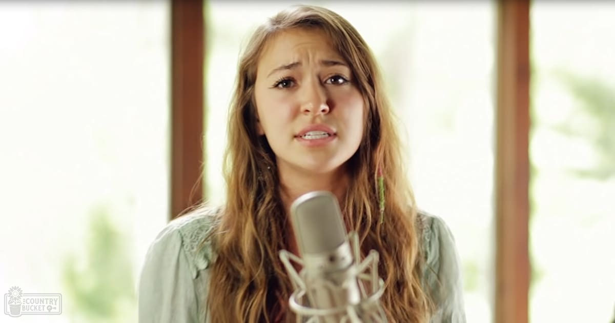 A young adult female wearing a light green blouse with lace detailing sings into a microphone in an indoor setting with natural daylight streaming through large windows.