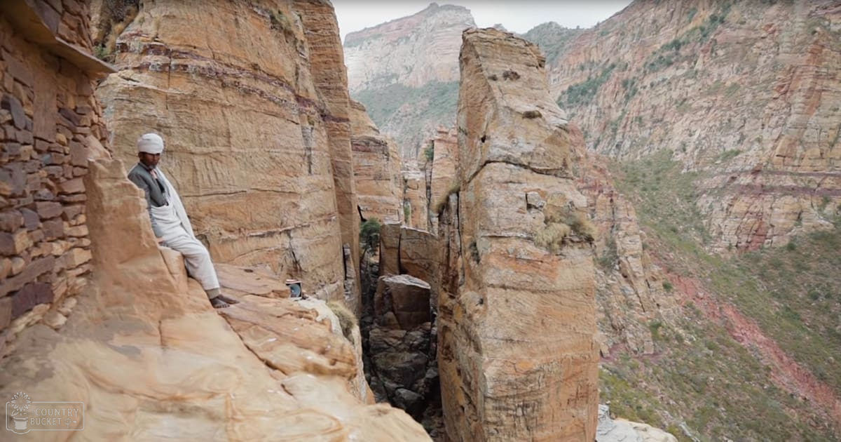 An adult male wearing a white turban, light-colored pants, and a light gray jacket stands in a rugged canyon with sparse vegetation under diffused daylight.
