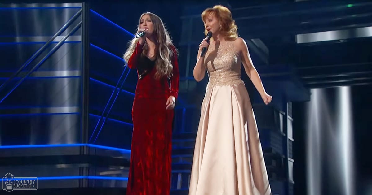 Two adult female singers perform on a stage with microphones and red and beige dresses in an indoor concert hall setting.