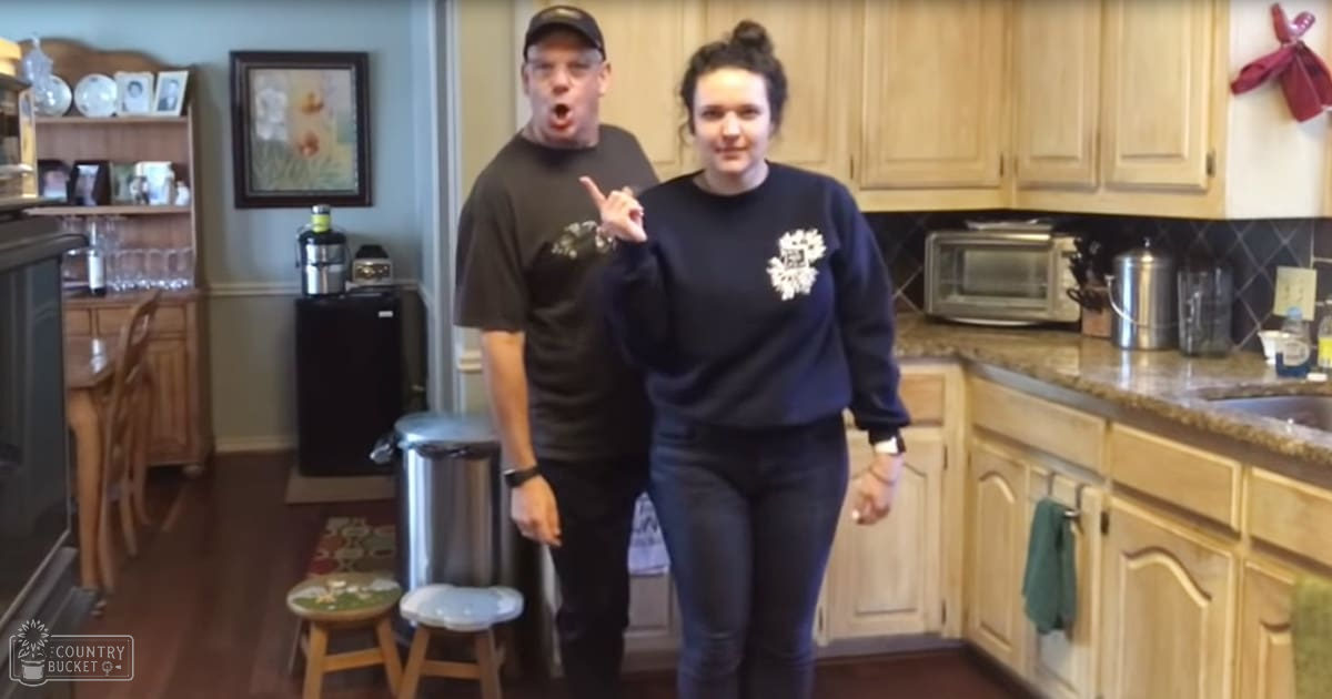 Two young adults in a well-furnished kitchen with wooden furniture and natural light, one wearing a black t-shirt and the other a dark blue sweatshirt with white design.
