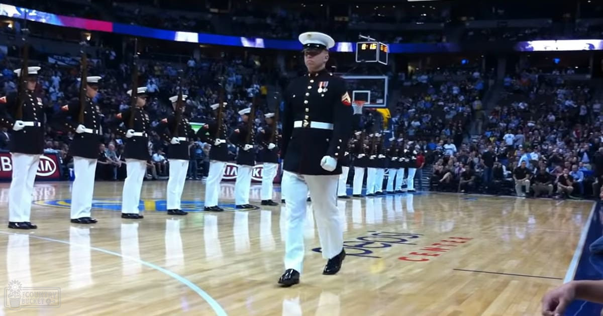Adult male military personnel in a dark blue dress uniform, white gloves, and black shoes standing at an indoor sports arena with markings typical of a basketball court.