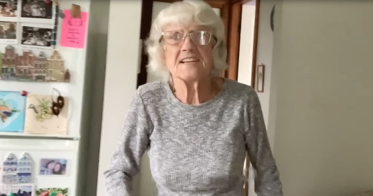 An older adult female wearing a gray ribbed sweater indoors, surrounded by household items like a refrigerator with magnets.