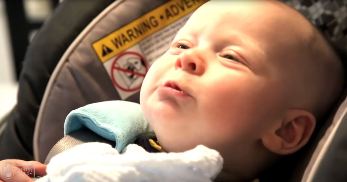 An infant in a white onesie with light blue collar, seated in a car seat indoors under soft natural light.