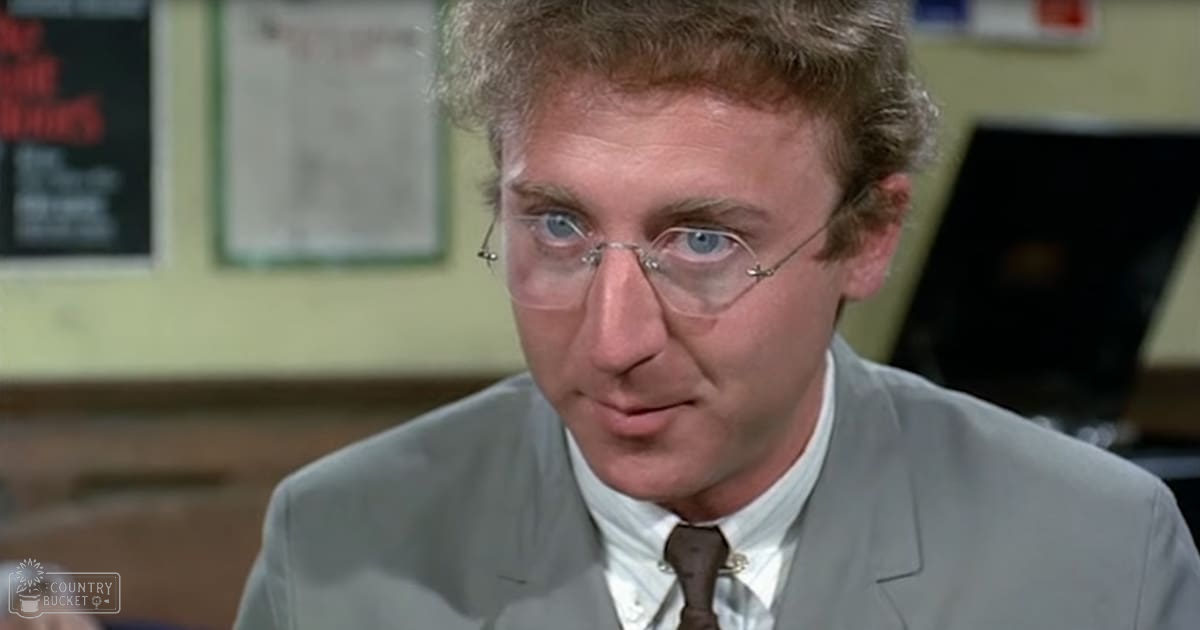Middle-aged adult male wearing a tailored light gray suit jacket, white dress shirt, and dark brown tie in an indoor office setting with papers pinned to a board behind him.
