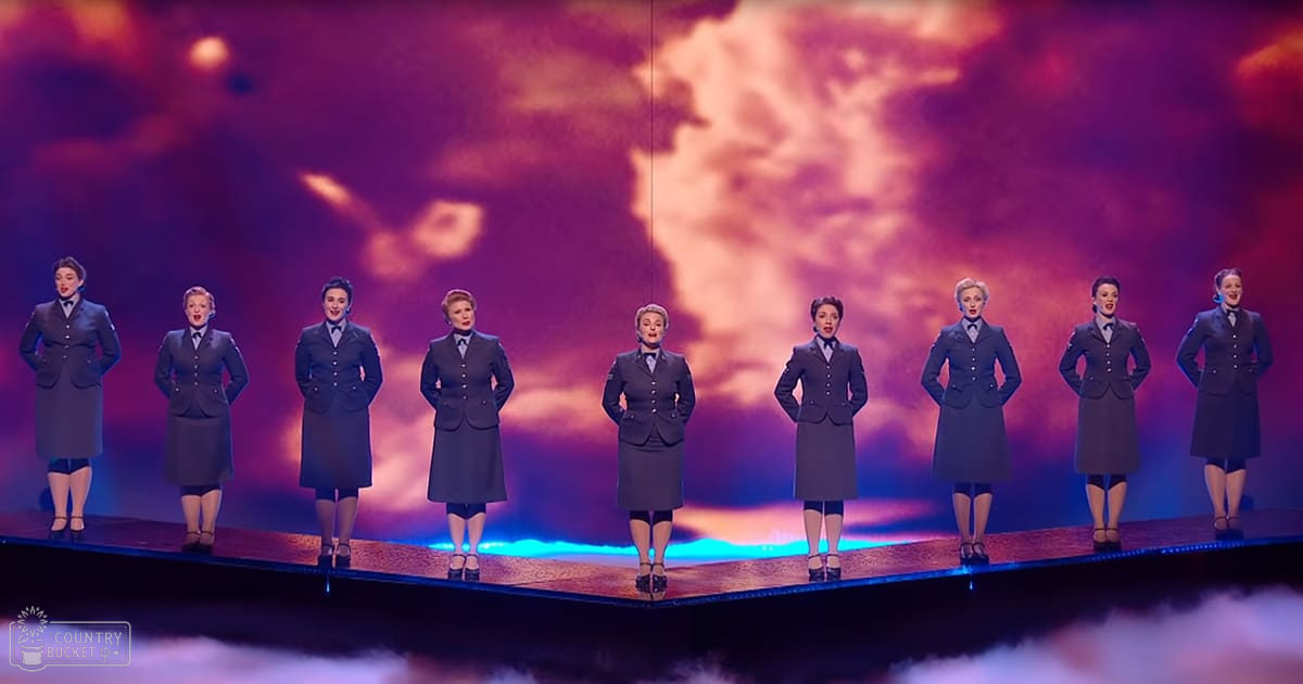 A theatrical performance on a stage with adult female performers in dark double-breasted jackets against a vibrant sky backdrop.