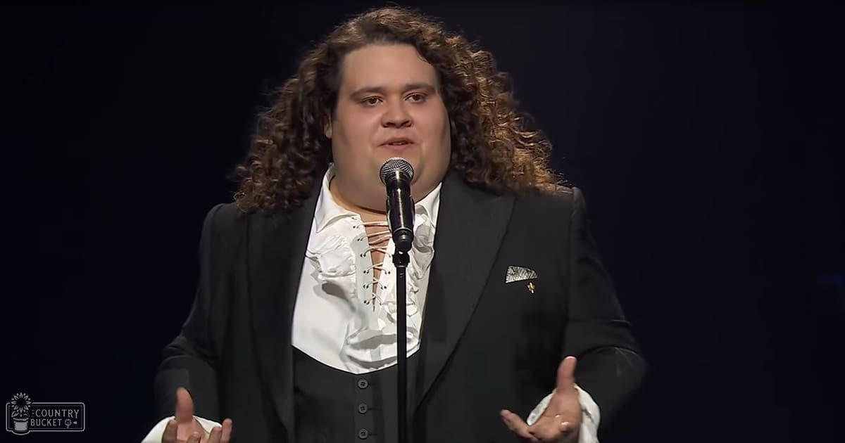 A man in an adult age band wearing a formal suit with a cravat-style tie stands at a microphone stand indoors, suggesting a speech or presentation setting.