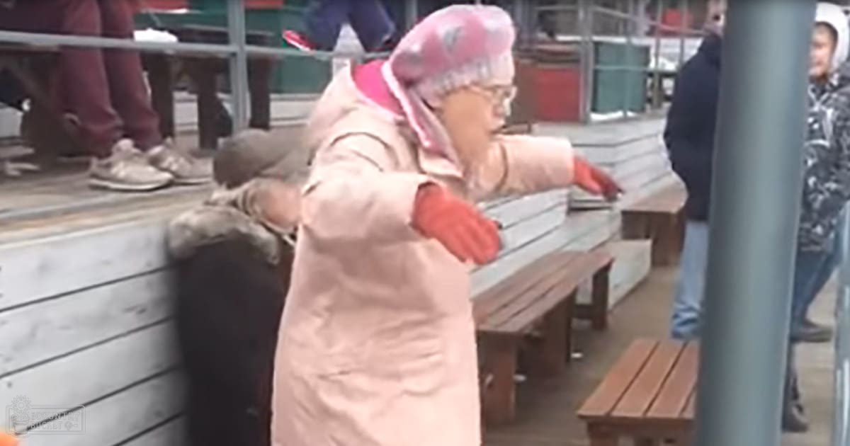 An older adult female wearing a light pink coat and red gloves, engaged with her hands in an outdoor market setting with wooden benches and green containers in the background.