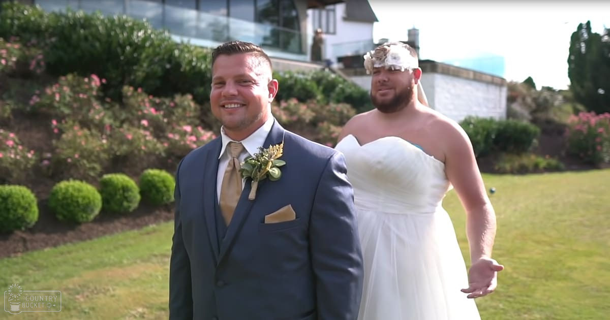 A wedding couple, a male in a dark blue formal suit with a light brown tie and pocket square, and a female in a strapless white wedding gown with a floral headpiece, standing outdoors at what appears to be a wedding venue.