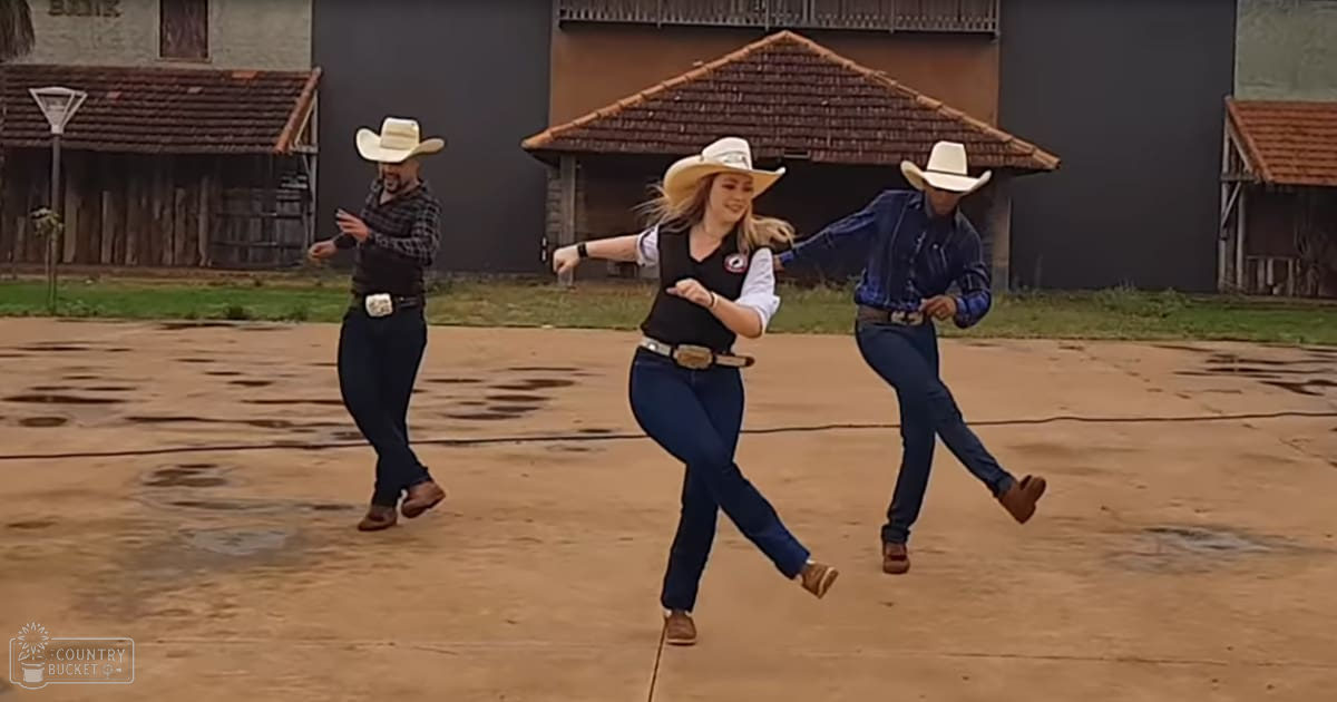 Three adults dancing outdoors near rural buildings with tiled roofs; one wears plaid shirt and cowboy hat, another in black vest over white shirt, both wearing brown shoes and large belt buckles.