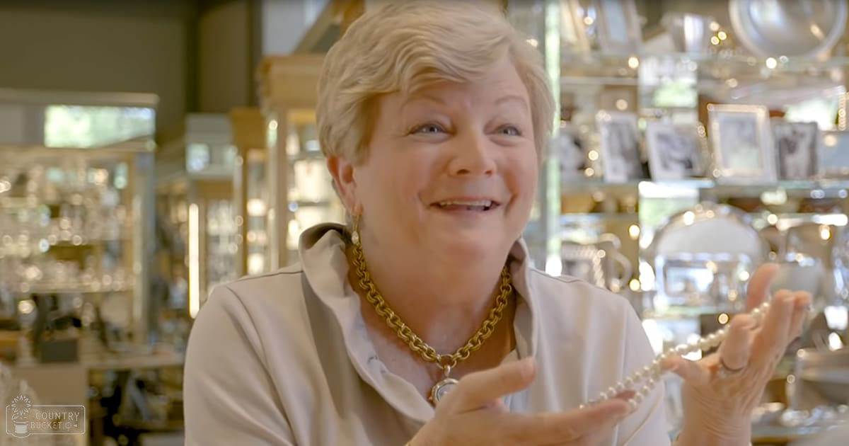 An older adult female in a jewelry store, wearing a beige collared shirt with a gold chain-link necklace and watch, holding a pearl necklace.
