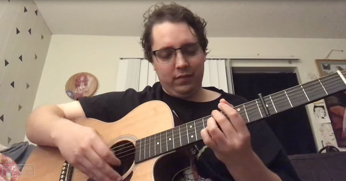 A young adult male plays a guitar in an indoor setting with geometric patterns on the walls.