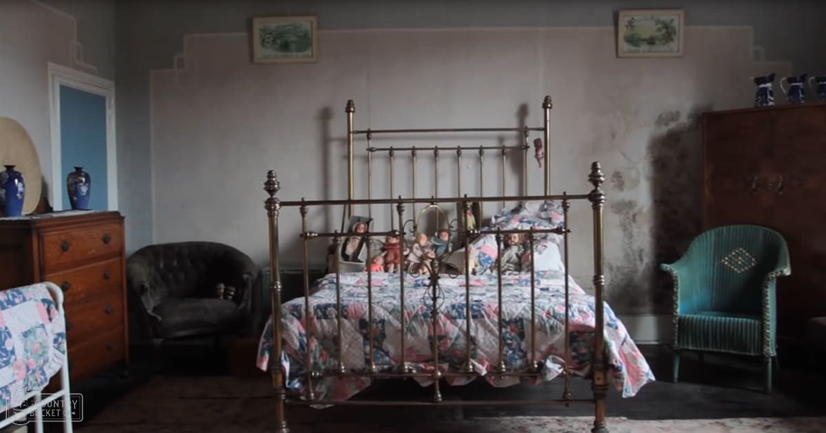 An older vintage bedroom scene with ornate brass bed, floral-patterned quilt, wooden dresser holding vases, dark leather armchair, teal upholstered chair, framed pictures above the bed, and muted tones of teal, beige, white, and shades of blue in wallpapered walls and a door leading out of the room