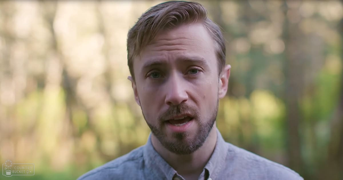 A young adult male wearing a modern casual button-down shirt with long sleeves stands outdoors amidst trees and greenery under soft diffused lighting.