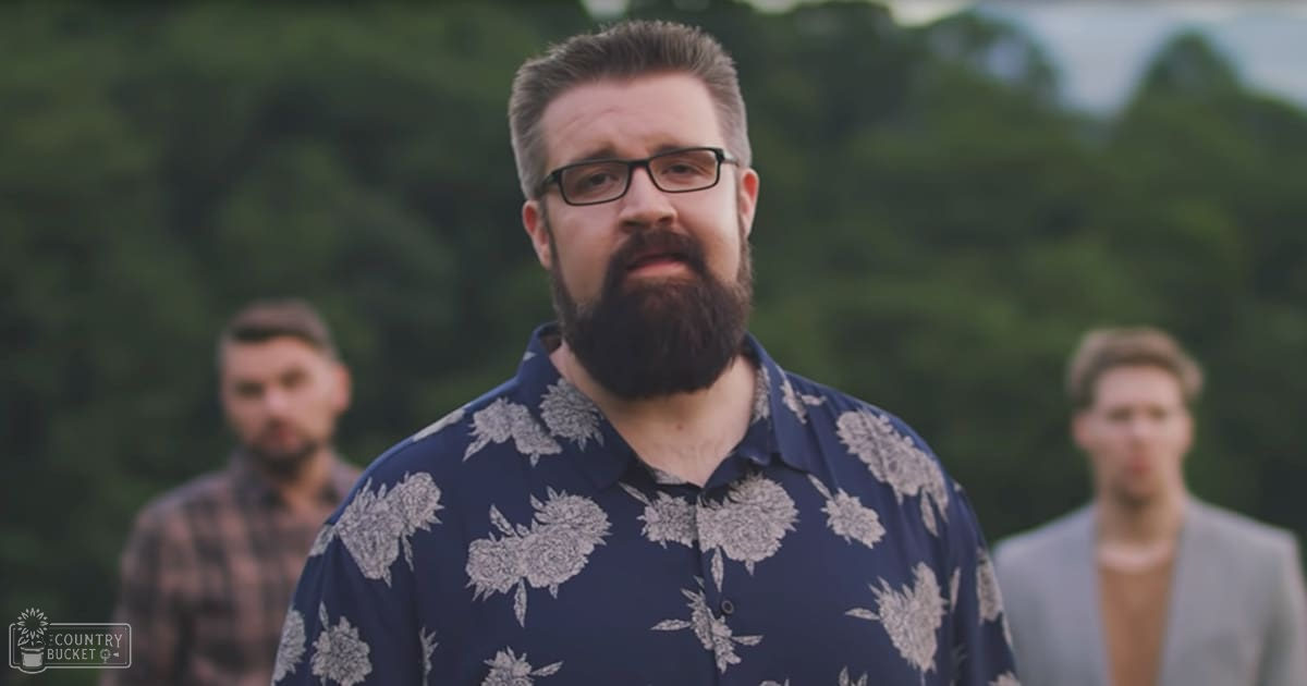A middle-aged man wearing a blue floral shirt stands outdoors with two young adult males in plaid shirts and light gray blazer respectively, surrounded by greenery under soft daylight.
