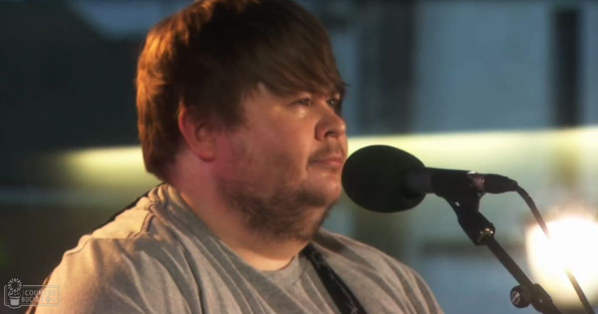 A middle-aged adult male wearing a gray, relaxed-fit crew-neck t-shirt with a white stripe across shoulders and back, standing in an indoor recording studio environment with professional audio equipment.