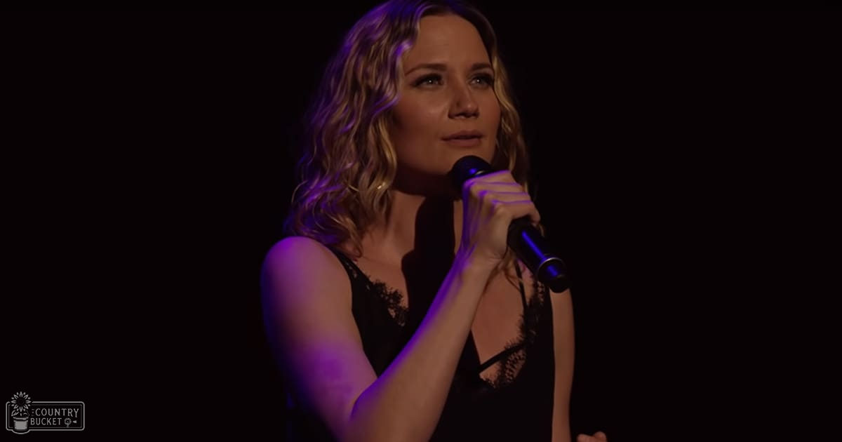 A young adult female in a black sleeveless lace top sings into a microphone at an indoor event with dim stage lighting.
