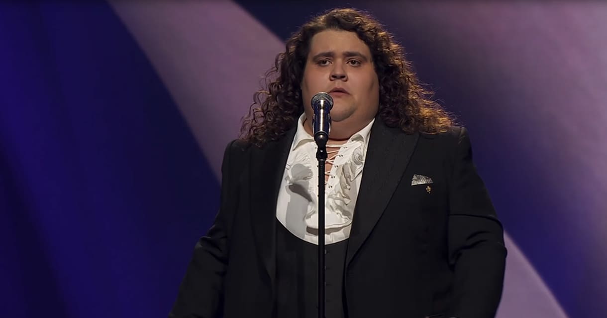 An adult male performer in a black suit with ruffled white shirt and purple stage lighting, likely during a concert or theatrical performance.