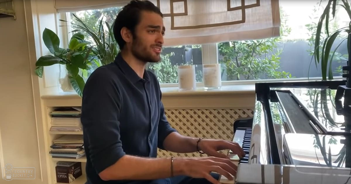 A young adult male plays a black grand piano in an indoor home setting with natural light and greenery.