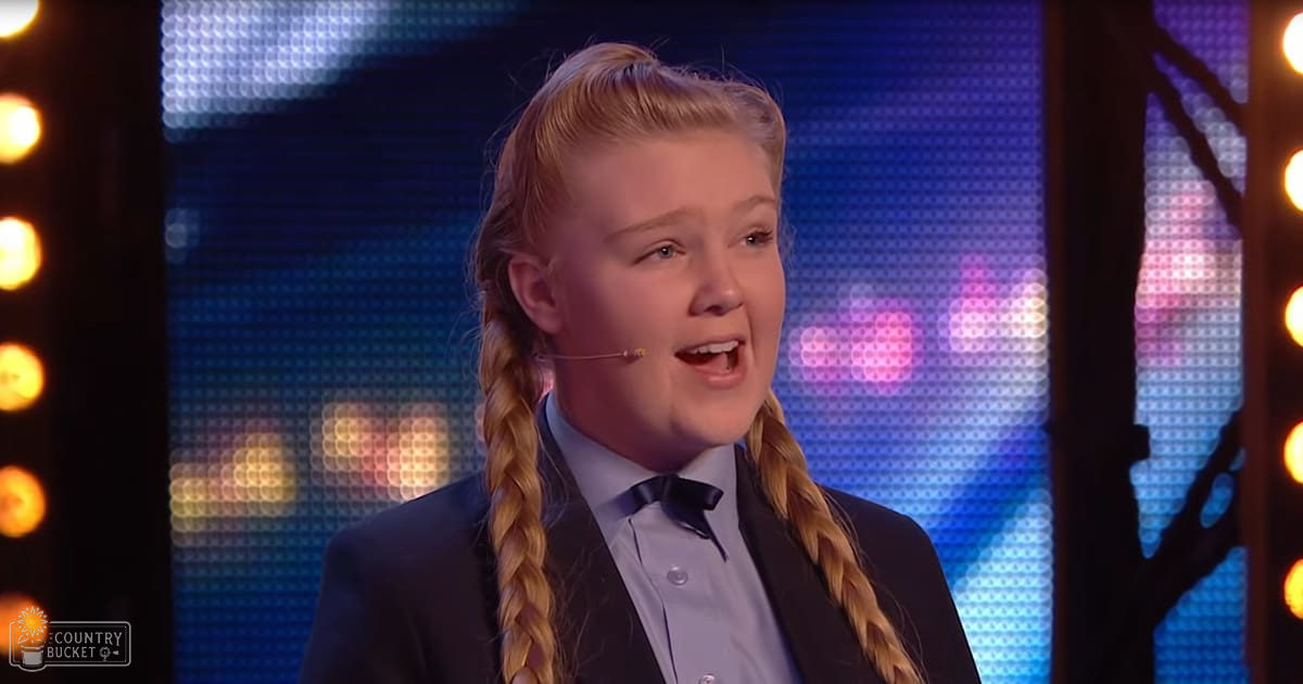 A young adult female with long blonde braids in a dark suit jacket over a light blue dress shirt and black bow tie, performing on stage at an evening talent show or competition.