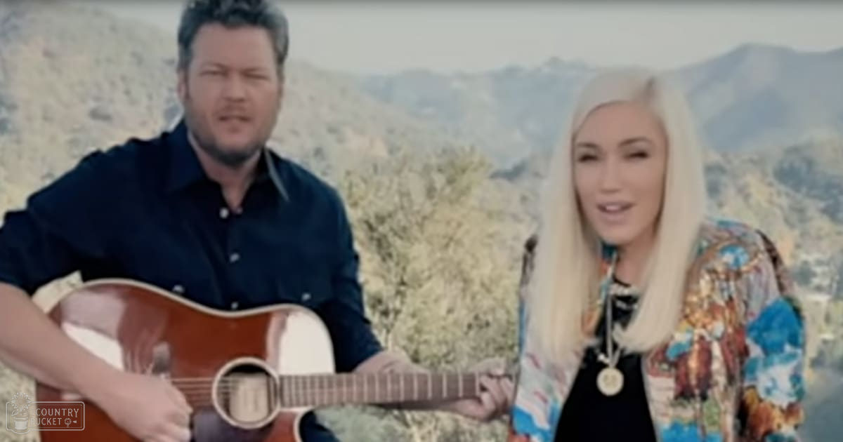 A middle-aged man plays an acoustic guitar outdoors while a young adult to middle-aged woman stands beside him wearing a colorful patterned jacket over a dark top, layered necklaces, and holding a gold-toned necklace. The setting is mountainous with lush greenery in late spring or summer under clear midday light.