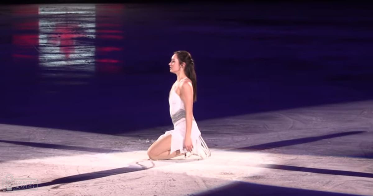 A young adult female in a sleeveless, high-necked white dress with light fabric stands on an indoor ice rink under artificial lighting.