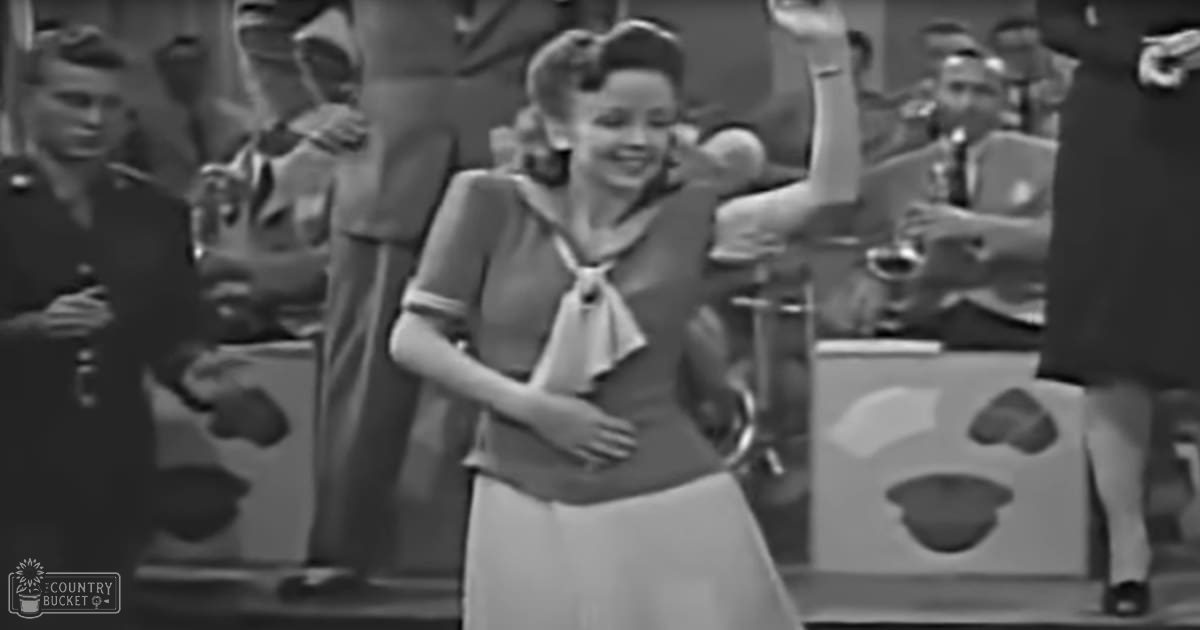 An indoor event or performance with a young girl in a white dress holding a small object, surrounded by adults dressed formally under artificial light during the day.