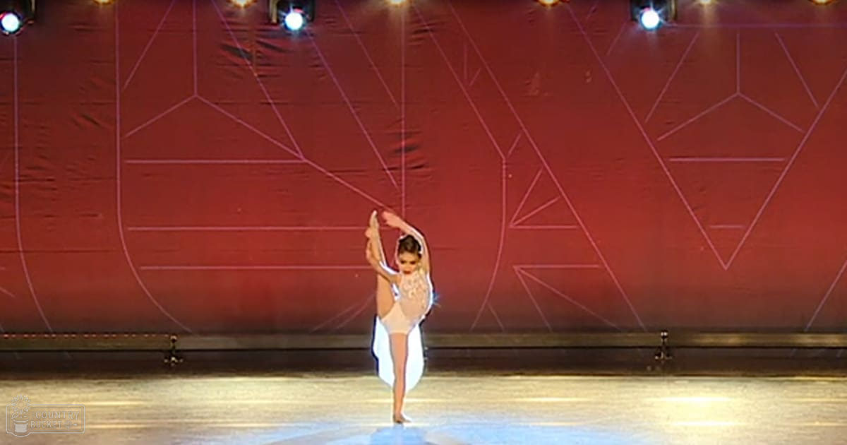 A young adult female dancer performing in a form-fitting, sleeveless white ballet leotard with a round neckline on an indoor stage with red geometric-patterned curtains and artificial stage lighting.