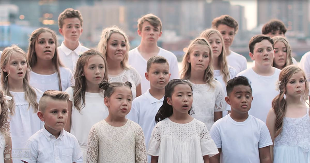 A group of children in a choir setting outdoors with a cityscape background. The children appear to be between 6-18 years old, predominantly dressed in white shirts and dresses with lace details or floral patterns.