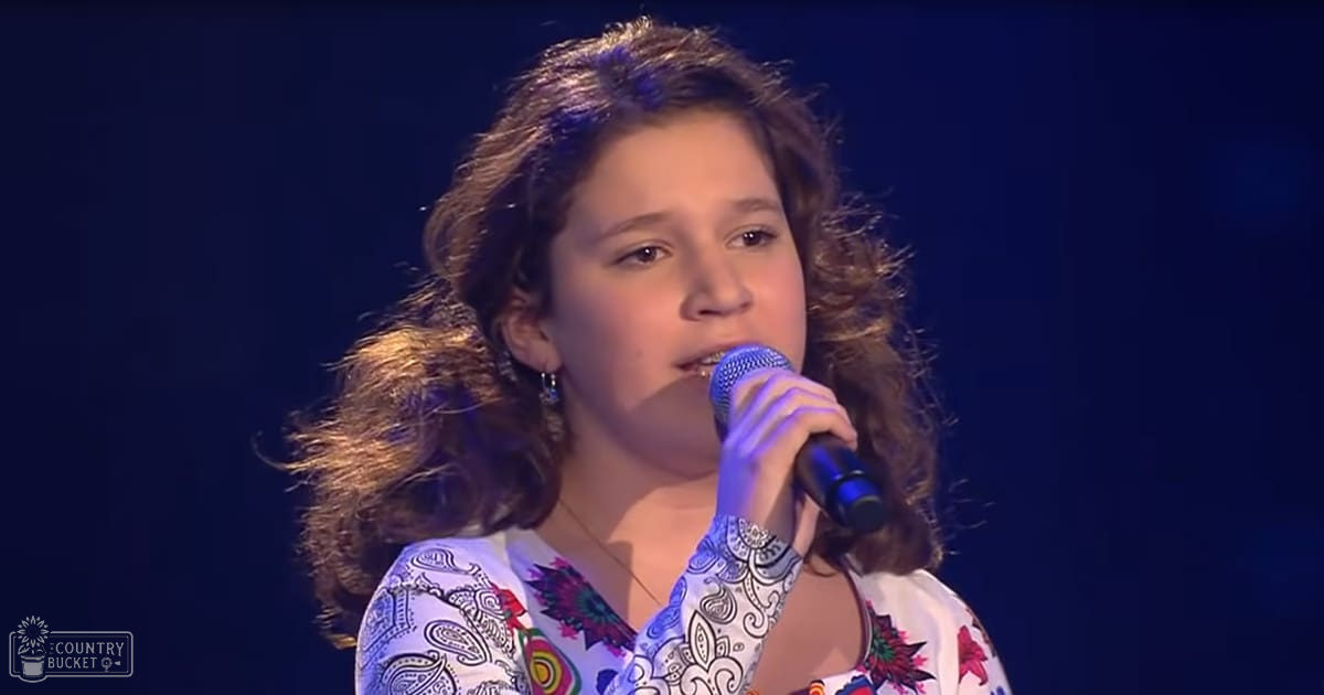A young adult female in a loose-fitting, long-sleeved floral and paisley-patterned top performs on stage under blue lighting.