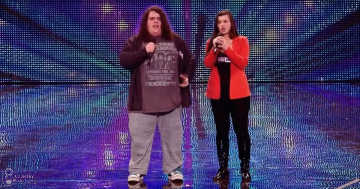 Young adult male singer in a long-sleeved graphic t-shirt with text and images, possibly band-related, and gray sweatpants or casual pants, alongside a young adult female in a red blazer or cardigan with black top underneath, black leggings or tights, and black boots, performing on stage with colorful digital backdrop displaying dots and lines.