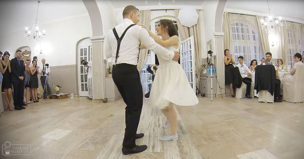 A wedding reception scene with a couple dancing under chandeliers and large windows draped with curtains.