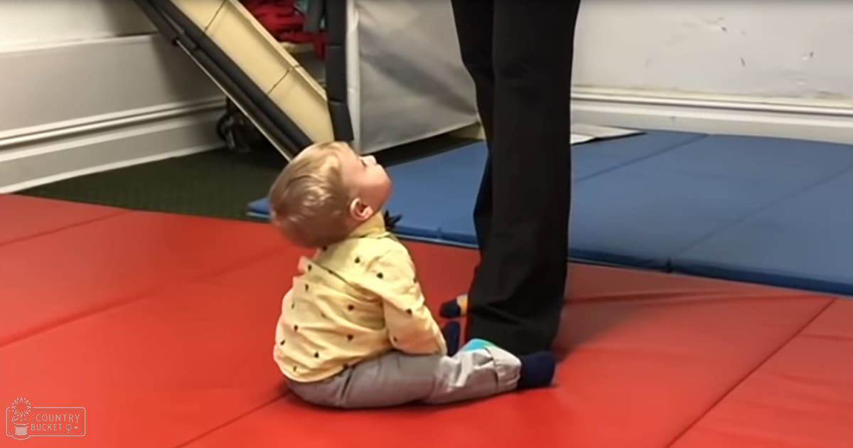A toddler wearing a yellow shirt with small black polka dots sits on a red mat indoors, surrounded by wearied white walls and blue mats.