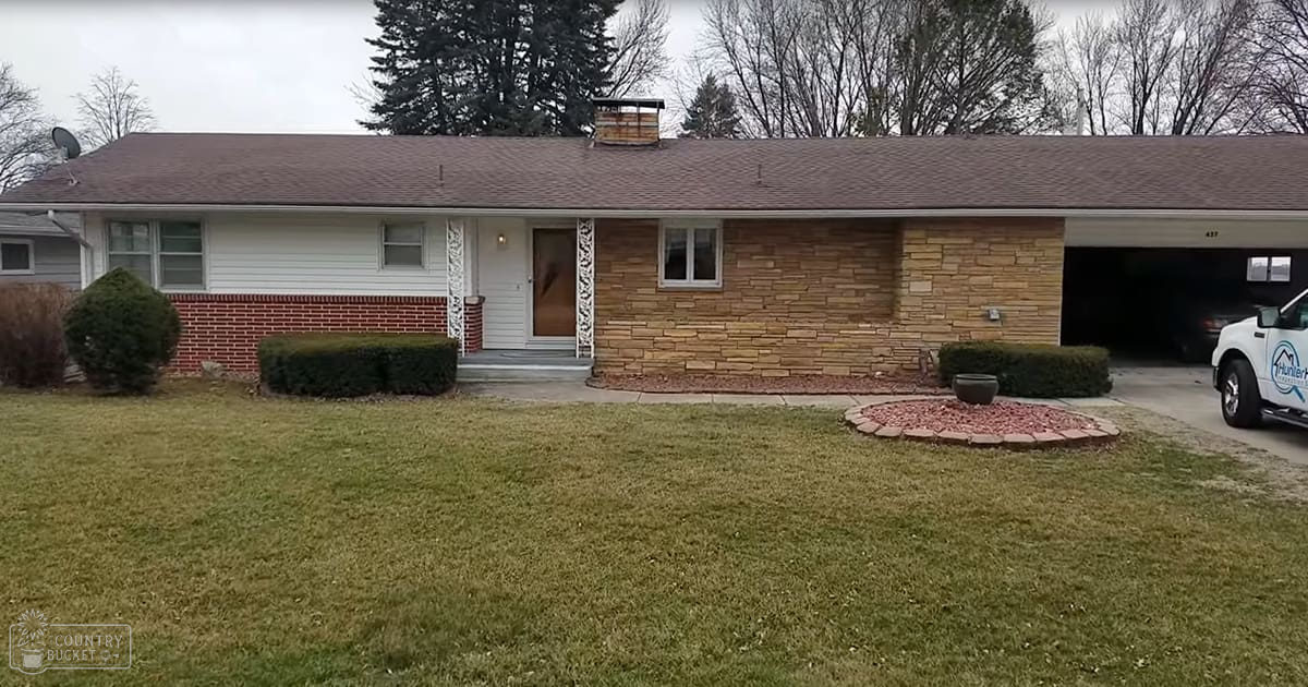 A suburban residential neighborhood with a white truck labeled 'Hunter', a small garden bed, and brick and stone houses. Trees suggest late autumn or early spring, overcast daylight.
