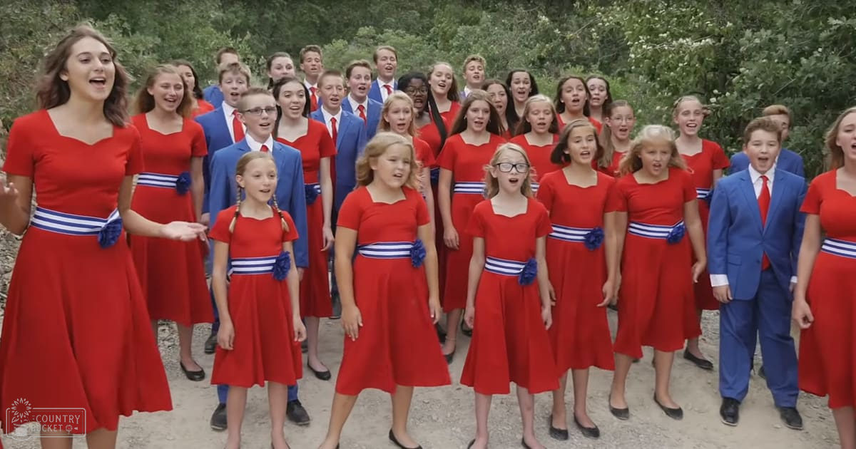 Children's choir performing outdoors with red dresses and blue suits amidst greenery in a natural setting, likely spring or summer.