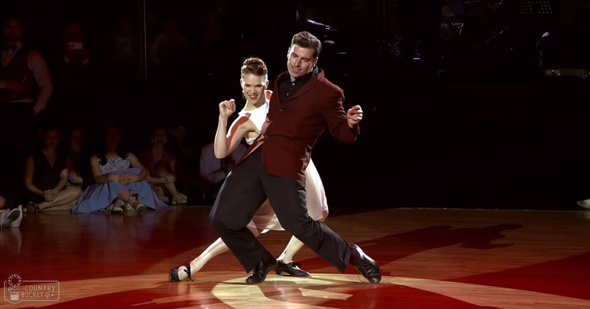 Two dancers in formal attire perform in an indoor ballroom setting, surrounded by spectators seated on benches and chairs.