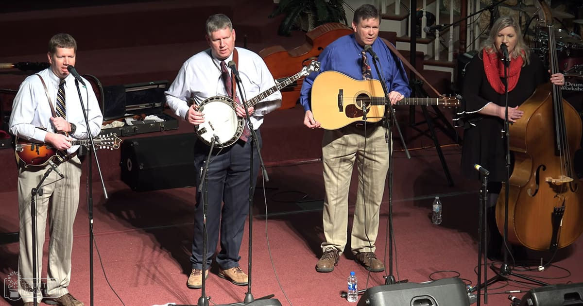 Adult male singer in light-colored pants, white striped shirt with banjo strap; middle-aged man holding a banjo, wearing dark blue pants, white shirt with red tie; adult male playing acoustic guitar in khaki pants and blue long-sleeve shirt; adult female in black dress with red scarf holding double bass. Performance venue or church setting with carpeted floors, plants, microphones on stands, natural light suggesting daytime.