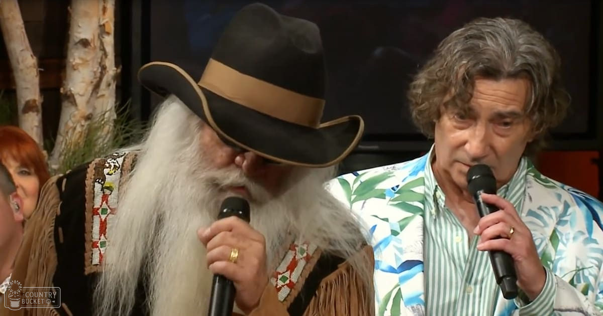 Two older adult males singing together indoors, one wearing a traditional Native American-style outfit with beadwork and fringe details on the shoulders, another in a colorful tropical leaf-patterned shirt; tree trunks and greenery in the background suggest an indoor setting themed around nature.