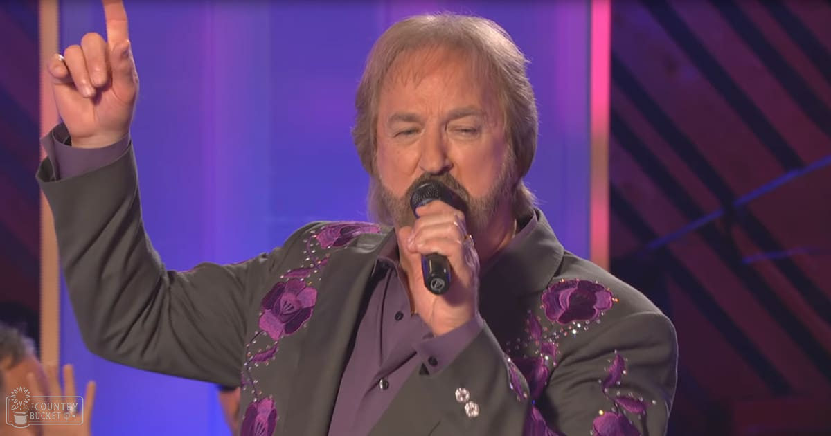 A middle-aged man performing on stage, wearing a dark gray suit jacket with purple floral embroidery and rhinestones, holding a microphone in his right hand under indoor performance lighting.