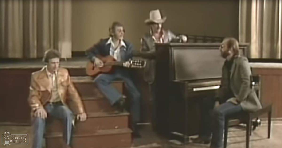 Musical performance in an indoor setting; adult males wearing casual attire seated around a piano under dim lighting.