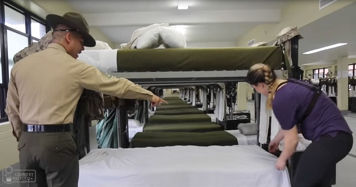 Adult male in a military training facility, wearing a tan uniform shirt with dark green pants and a black belt; adult female adjusting a bed nearby. The scene includes bunk beds with green mattresses and white sheets under fluorescent lighting.
