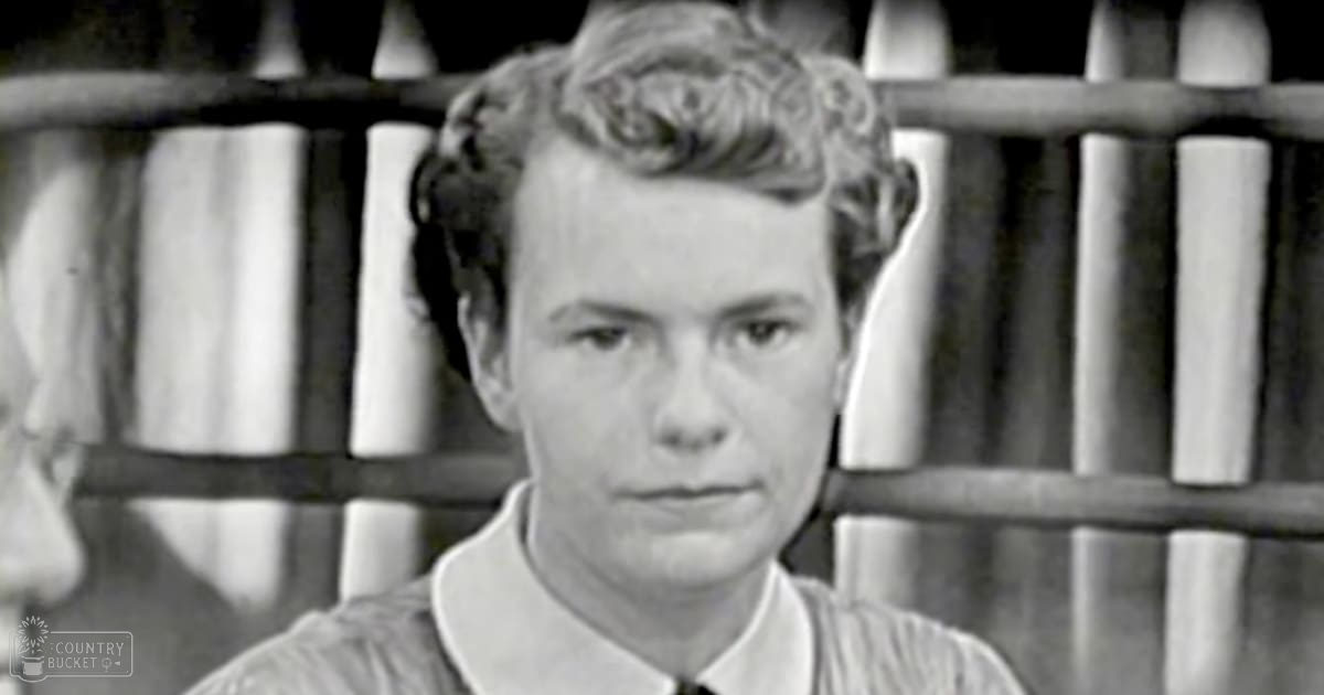 A young adult male wearing a mid-20th century collared shirt with a tie stands in the foreground while an older adult male in formal attire from the same era is partially visible in the background. The setting appears to be rustic, possibly outdoors or within a wooden structure.