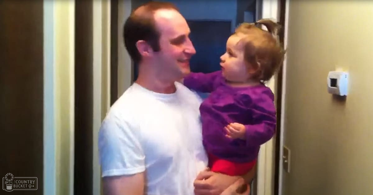 An adult male holding a toddler indoors, with the man wearing a white t-shirt and the child in a purple ruffled top and red pants. The setting appears to be a hallway or entryway during daylight.