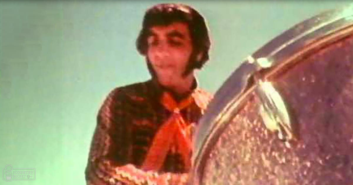 A young adult male wearing a patterned shirt with an orange tie, standing indoors against a light green wall.