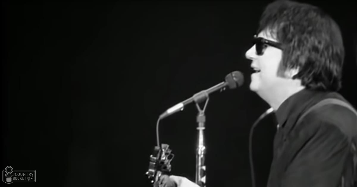 Adult male singer wearing a dark suit jacket and collared shirt, holding a microphone with an acoustic guitar in the background, set against a simple black backdrop.