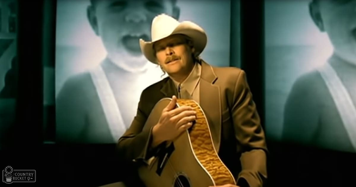 A middle-aged male singer wearing a tan suit jacket, white shirt, and cowboy hat holds a brown guitar strap against an indoor backdrop with blurred images of another person.