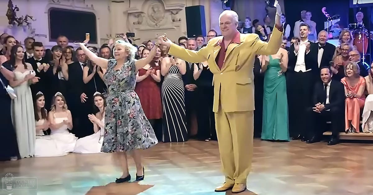 An adult male singer in a bright yellow suit dancing with an older adult woman in a floral dress at an elegant indoor event.