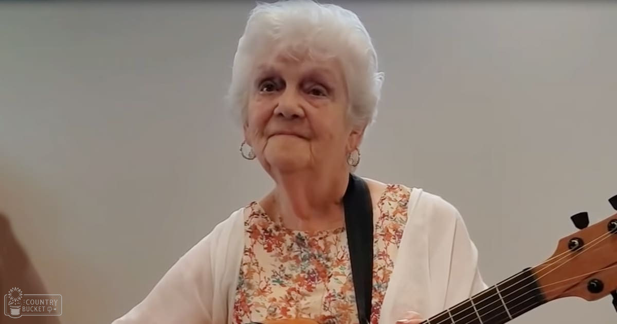 An older adult female wearing a floral-patterned top with a white cardigan or light jacket draped over her shoulders, holding a guitar strap around her neck in an indoor setting.