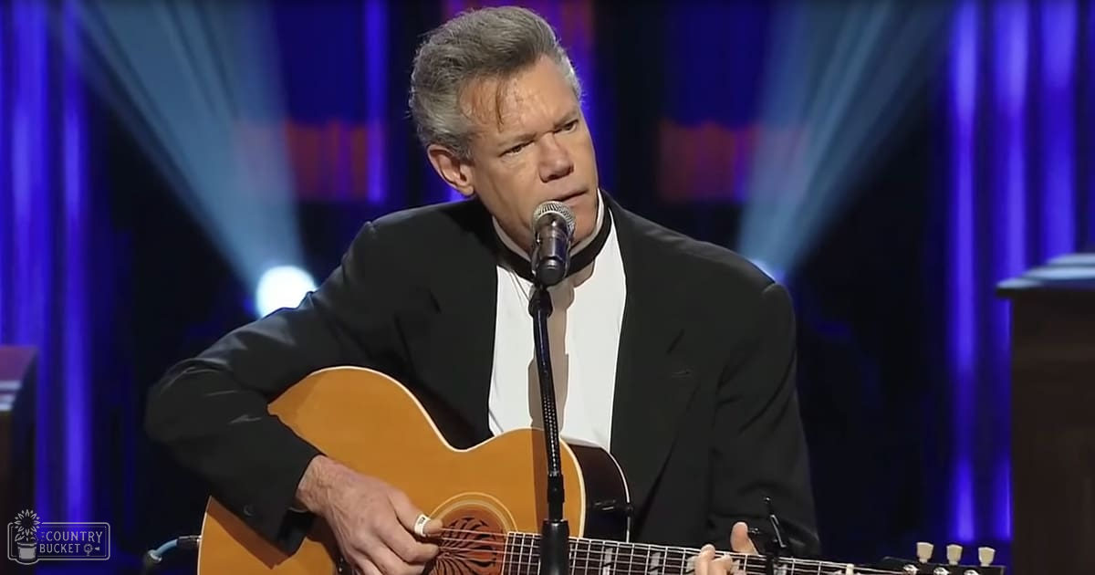 A middle-aged adult male singer in a black suit and white shirt performs on stage with an acoustic guitar, illuminated by spotlights under curtains at an evening concert.