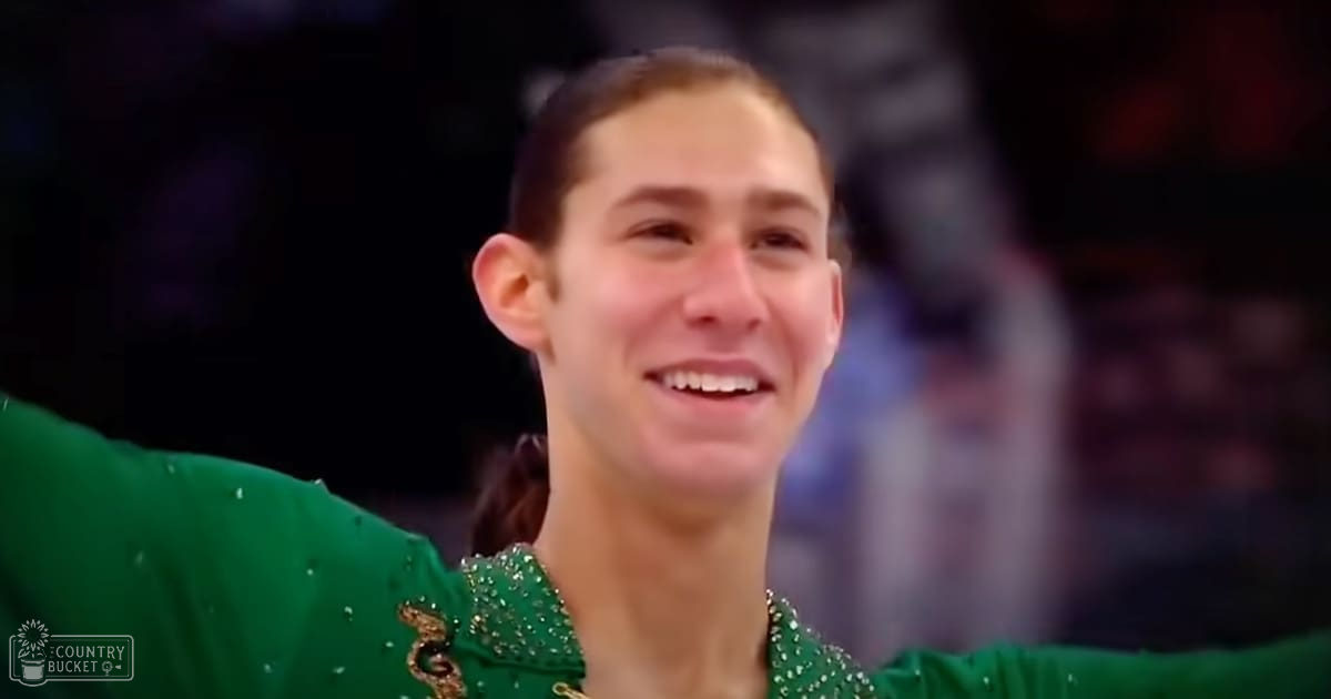 A young adult male with short brown hair wearing a green, shiny, figure skating costume in an indoor arena setting.
