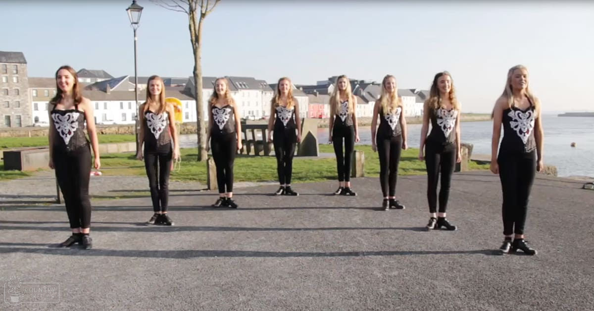 Group of young adult females standing by a waterfront, wearing black sleeveless tops with white floral embroidery and black leggings, on a paved area surrounded by grassy land and buildings in spring or summer.